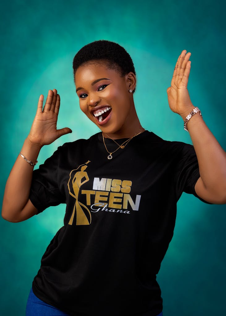Cheerful teenager posing energetically in a Miss Teen Ghana shirt against a teal background.